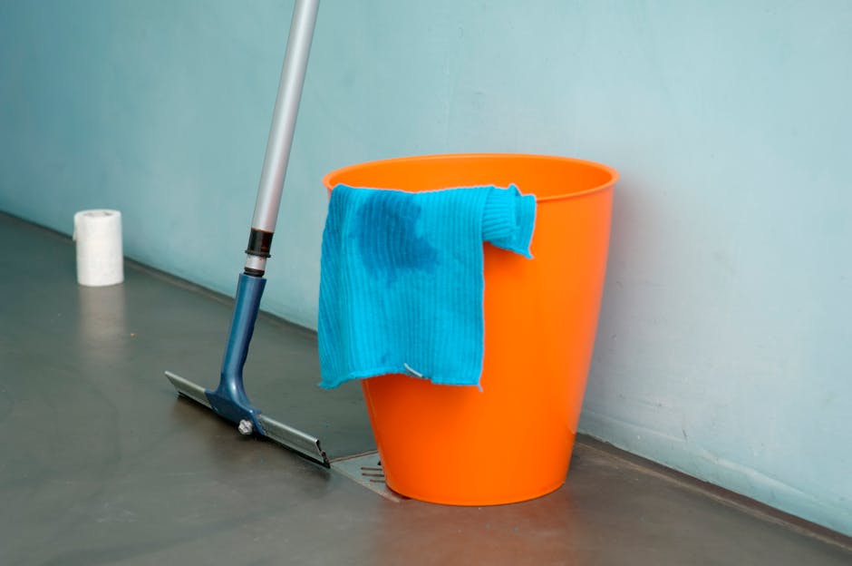 A bright orange plastic bucket with a blue cleaning cloth draped over its rim sits on a smooth, gray floor near a light green wall. An upright mop handle with a blue mop head is positioned nearby, par