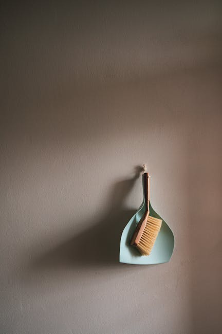 A bright orange plastic bucket with a blue cleaning cloth draped over its rim sits on a smooth, gray floor near a light green wall. An upright mop handle with a blue mop head is positioned nearby, par
